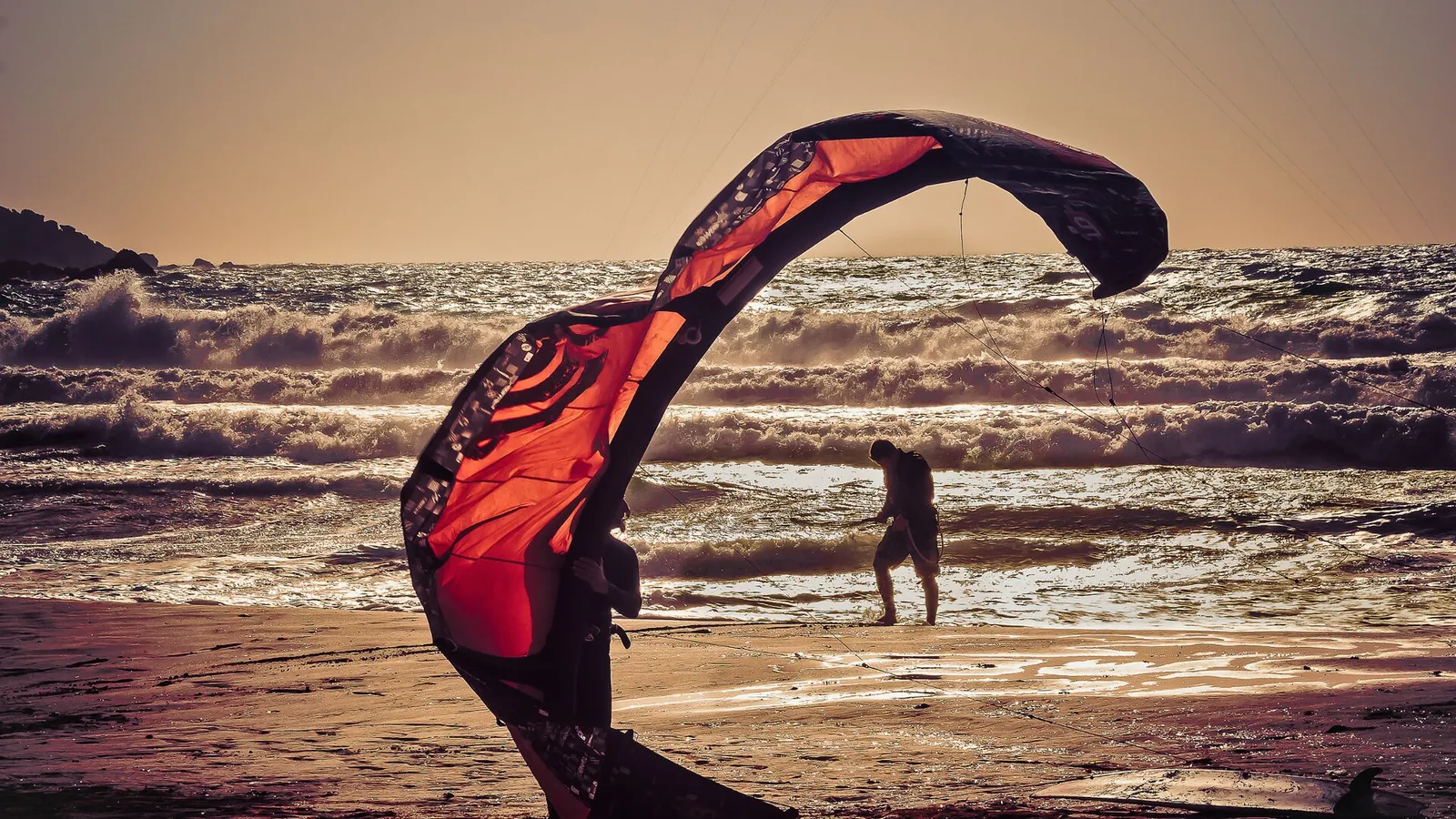 Two kitesurfers launching a kite on the beach at golden hour with breaking waves behind them