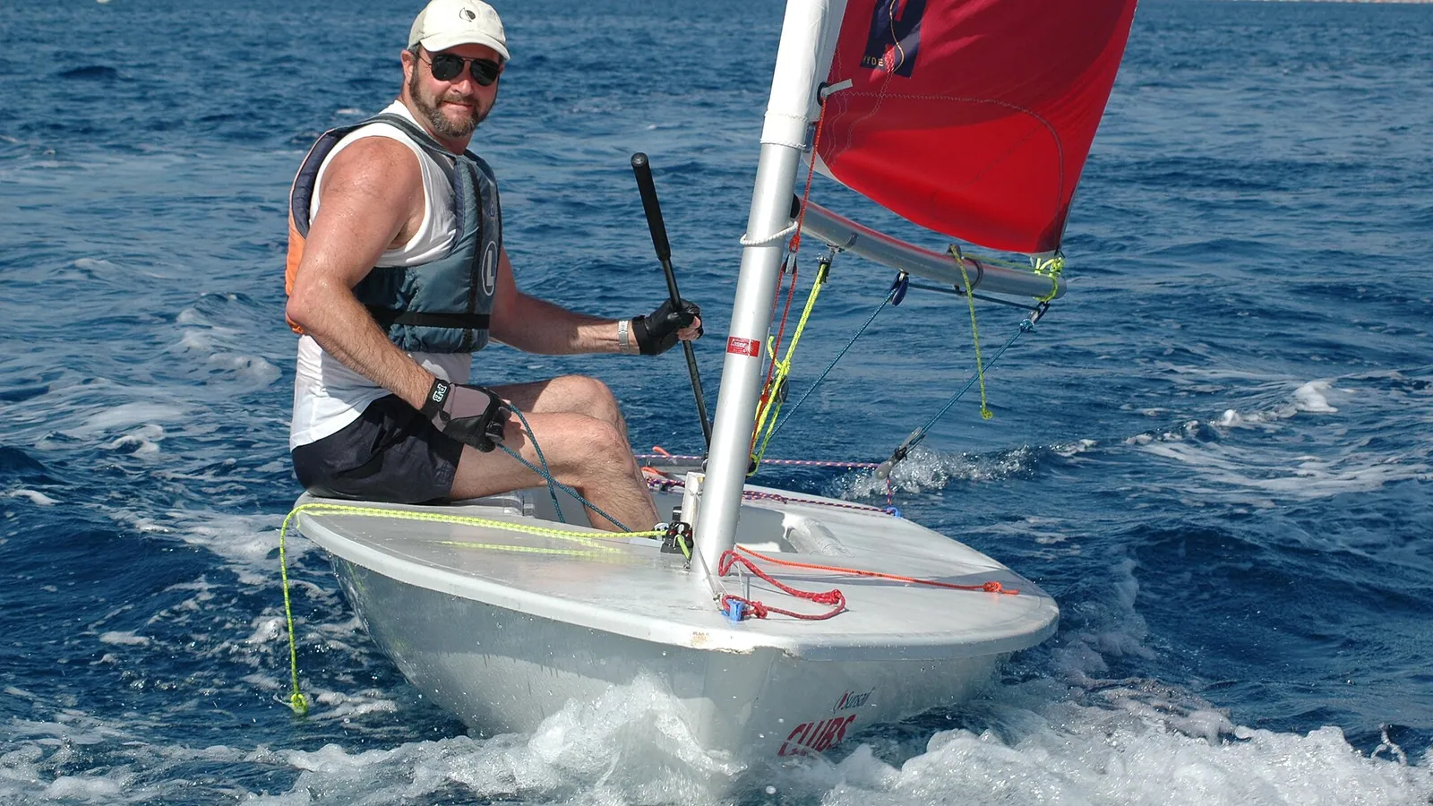 A Laser dinghy sailor planing across blue water in a fresh breeze