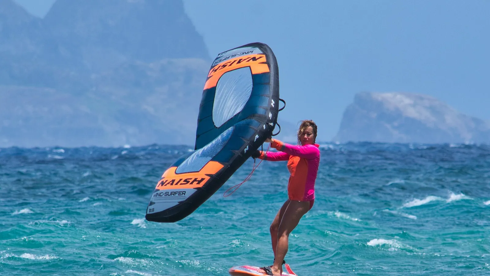 A wing foiler riding a hydrofoil board while holding a hand wing, mountains behind